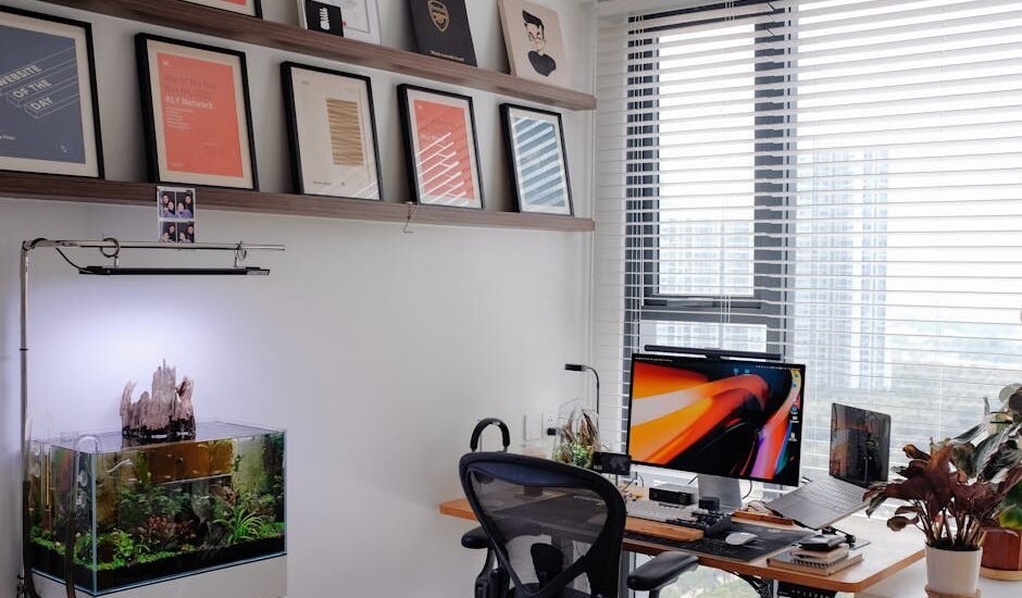 A planted aquarium with a visible surface skimmer sits beside a computer desk in a bright, modern home office setup.