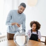 Man cleaning a round fish bowl with a net while a child watches, illustrating aquarium gravel vacuum care tips.