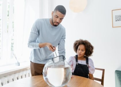 Man cleaning a round fish bowl with a net while a child watches, illustrating aquarium gravel vacuum care tips.