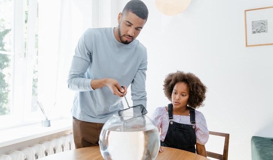 Man cleaning a round fish bowl with a net while a child watches, illustrating aquarium gravel vacuum care tips.